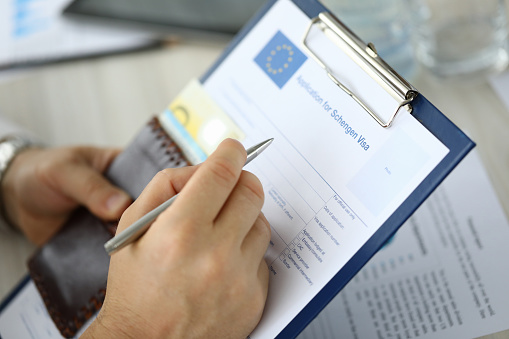 Close-up of person filling application form for schengen visa on clipboard. Man holding silver pen. Writing personal information on paper. Travelling abroad or immigration concept